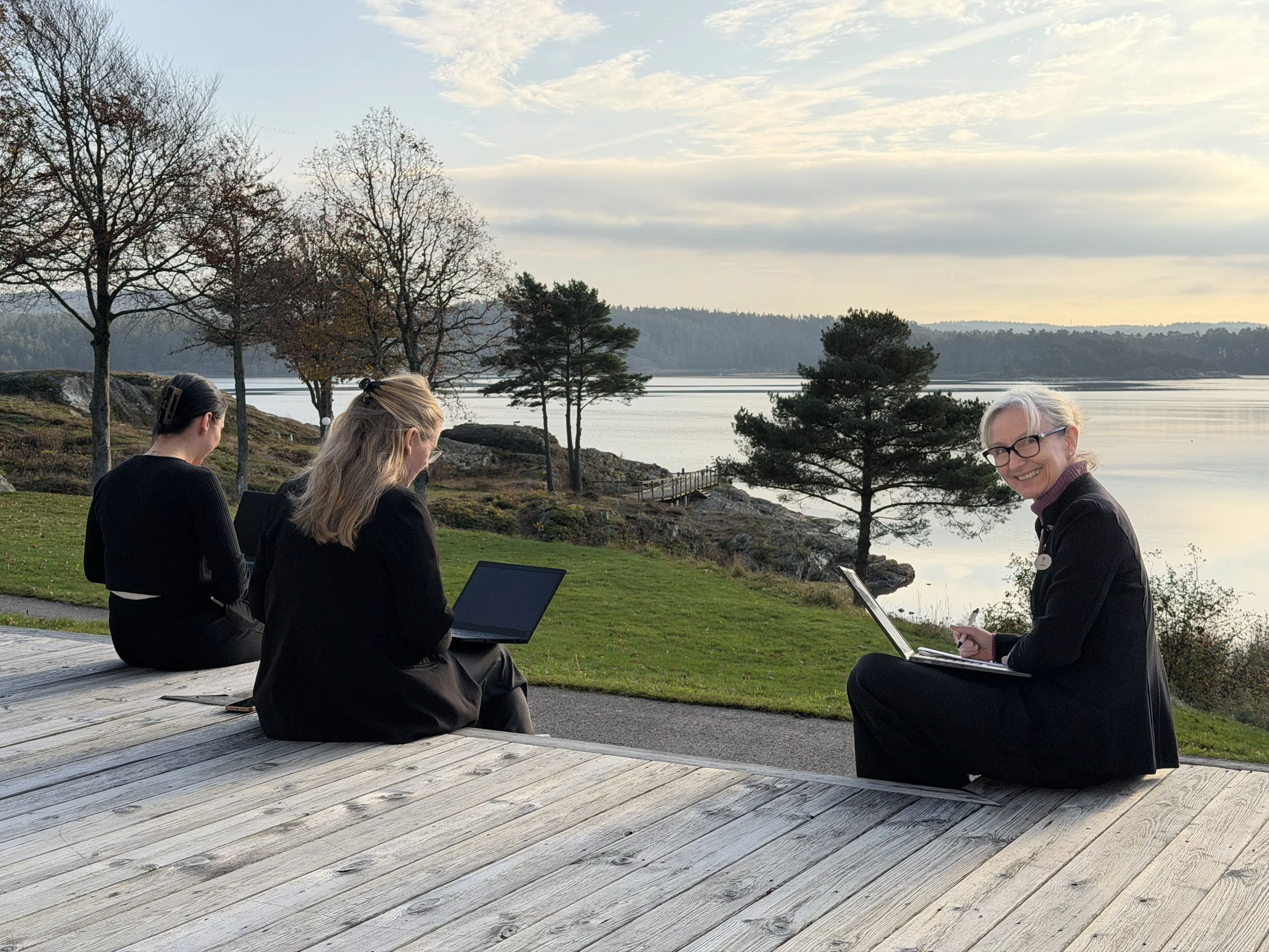 Tre personer som arbetar utomhus vid en fjord, alla tre med varsin laptop under en eftermiddag med soligt men lite disigt väder.