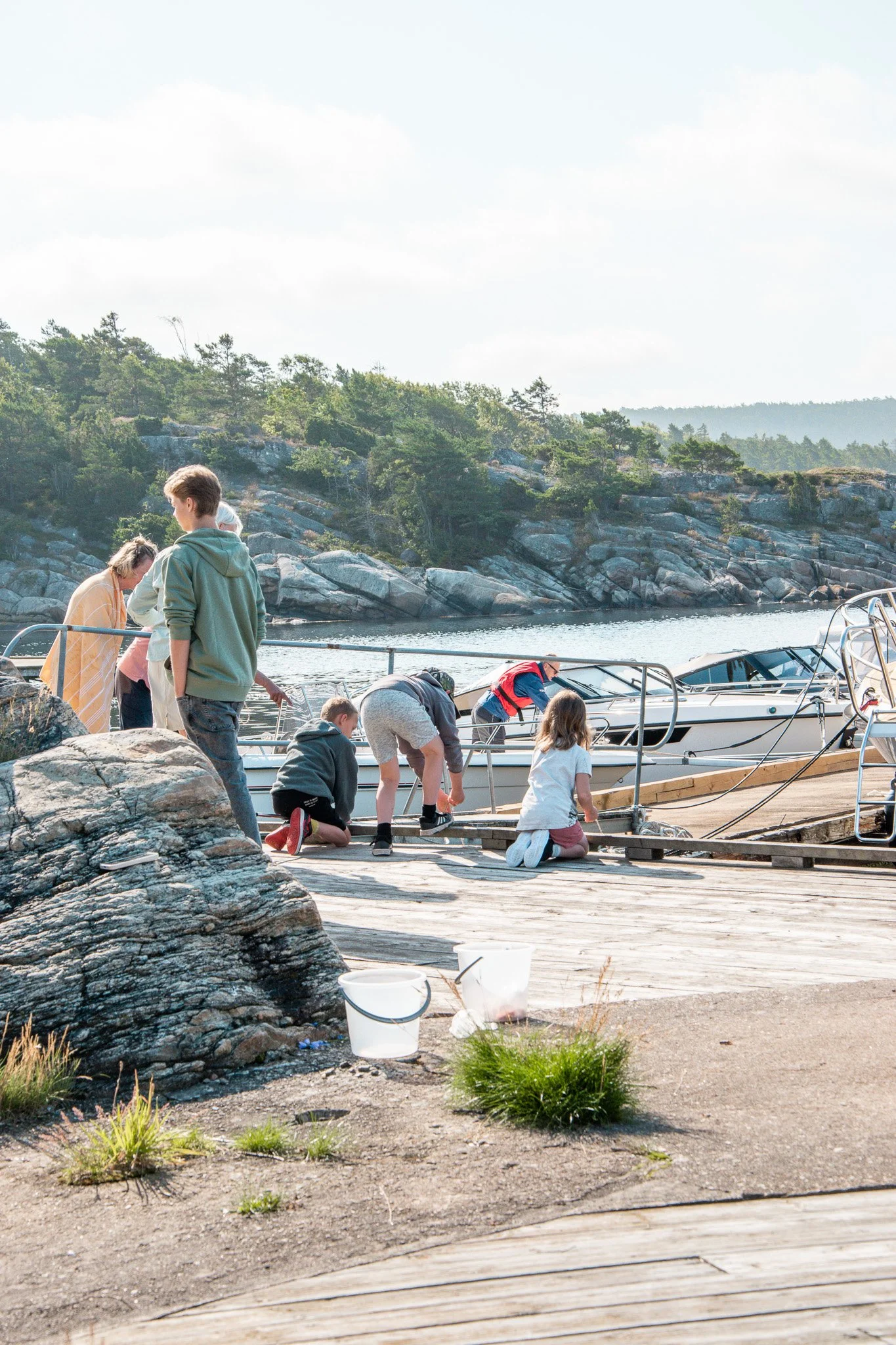 Flera barn och vuxna vid en brygga med båtar, omgivning av klippor och träd vid vattnet på en solig dag.