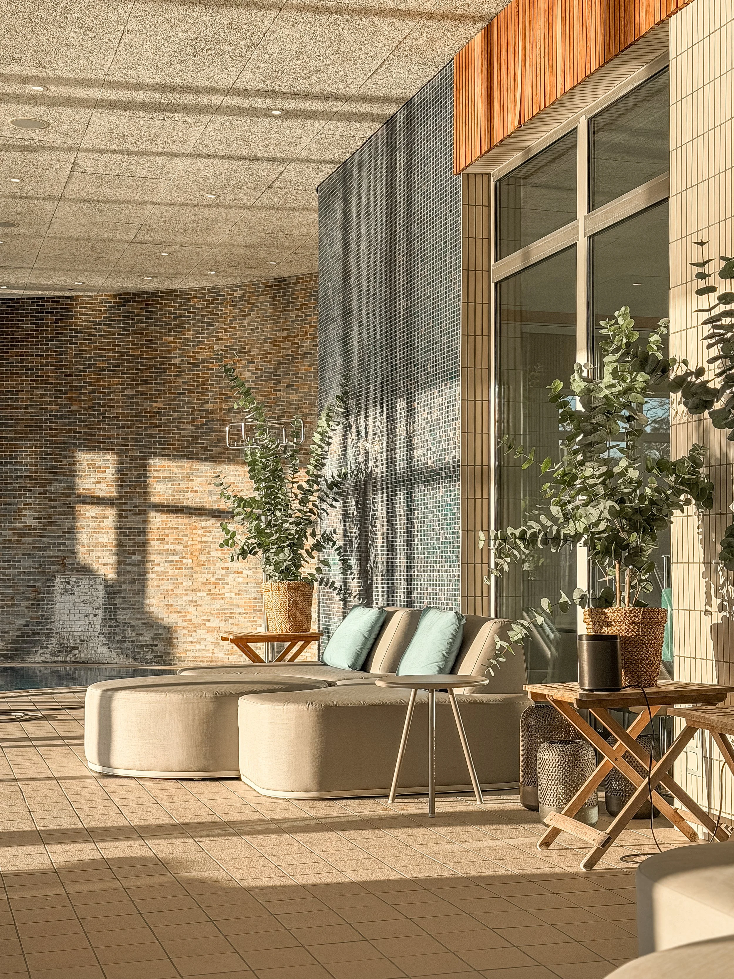 En solig loungeområde med ett runt vitt soffbord, bekväma fåtöljer med gröna kuddar, stora växter i mattade krukor, bruna tegelväggar och stora fönster som släpper in solljus.