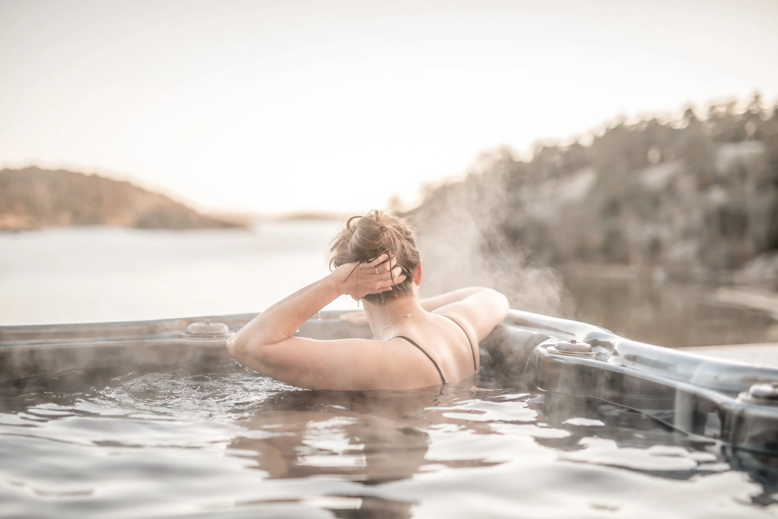 En person avkopplar i ett utomhus badkar på kvällen eller vid solnedgång, omgiven av natur och vatten.