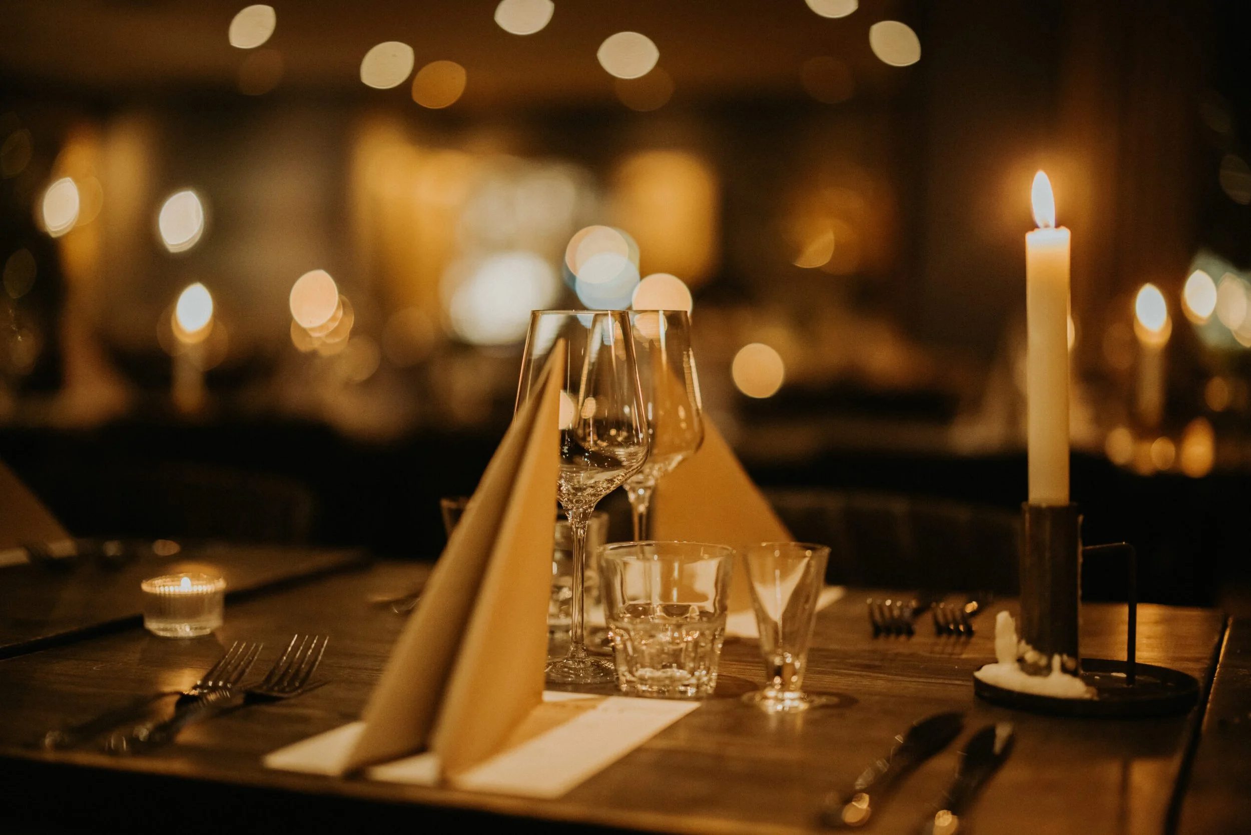 Ett dukat bord med tomma vinglas, bestick, en matta och ett lysande stearinljus i en restaurang med många bokeh-ljus i bakgrunden.