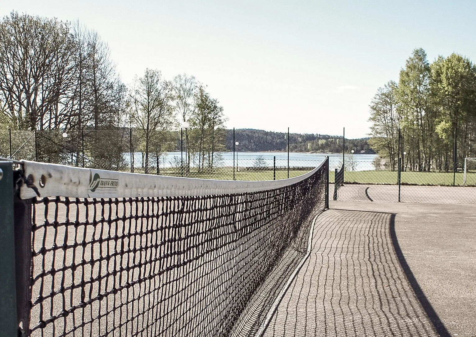 En tennisbana med nät och utsikt över en sjö och träd i bakgrunden under en klar, solig dag.