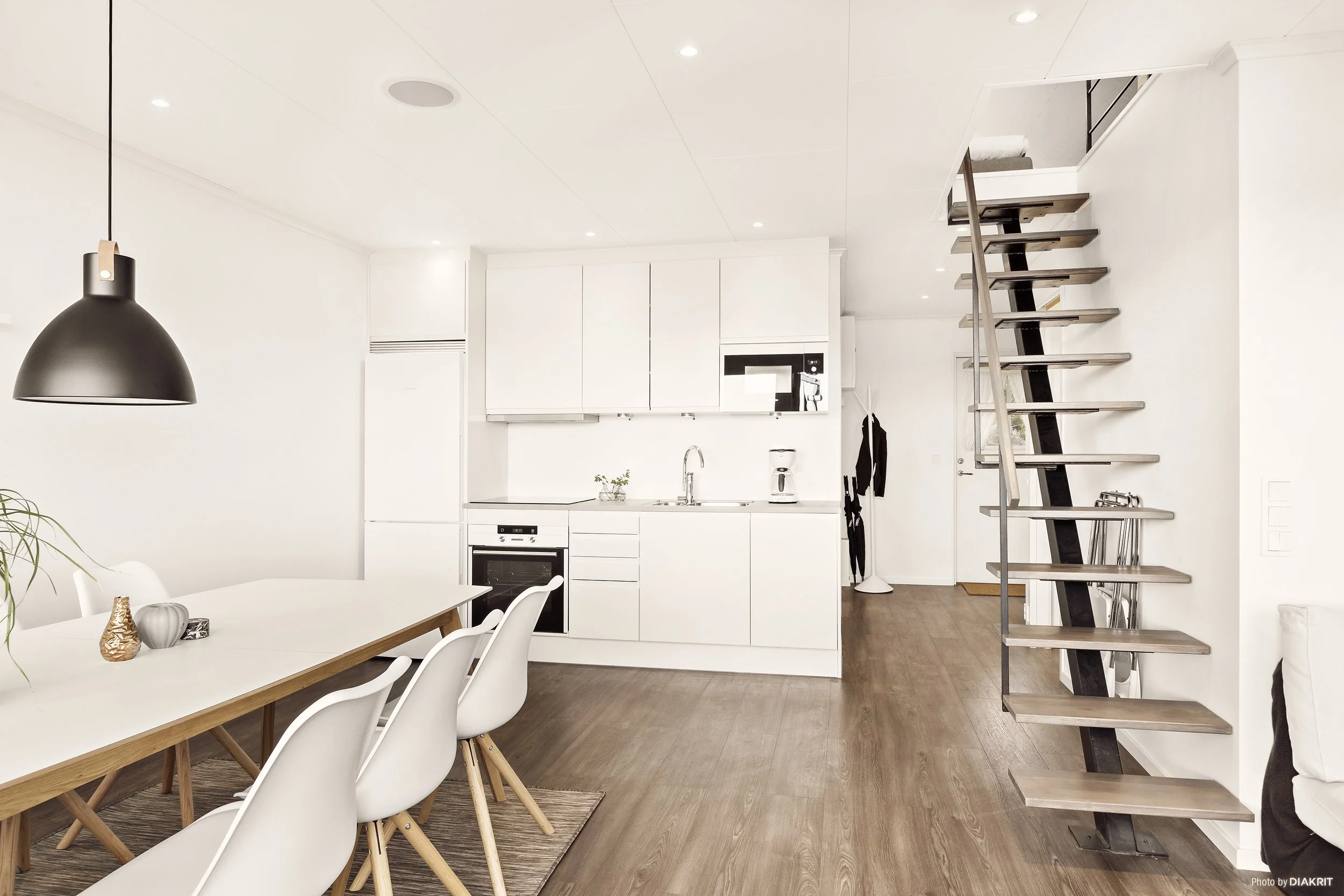 Ett modernt, minimalistiskt kök och matrum med vita väggar, träbänkar, vita stolar, ett svart taklampa, en köksö, och en öppen trapp framför en vit vägg.