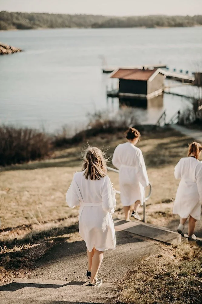 Tre kvinnor i vita sjukhusrockar går nerför en trappa mot en sjö, med hus och båtar i bakgrunden.