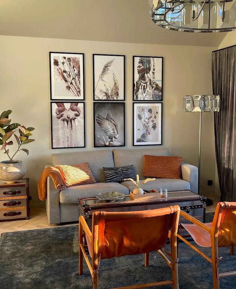 Vardagsrum med tavelvägg, beige soffa, två fåtöljer av trä och läder, böcker, lampor och växter.