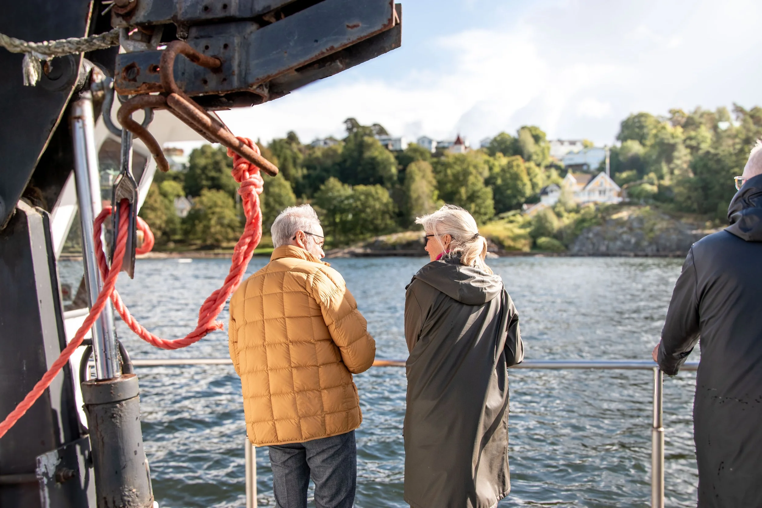 Tre personer står på en båt och tittar ut över vattnet med en landbacke med hus i bakgrunden, en dag med sol och lätt moln.