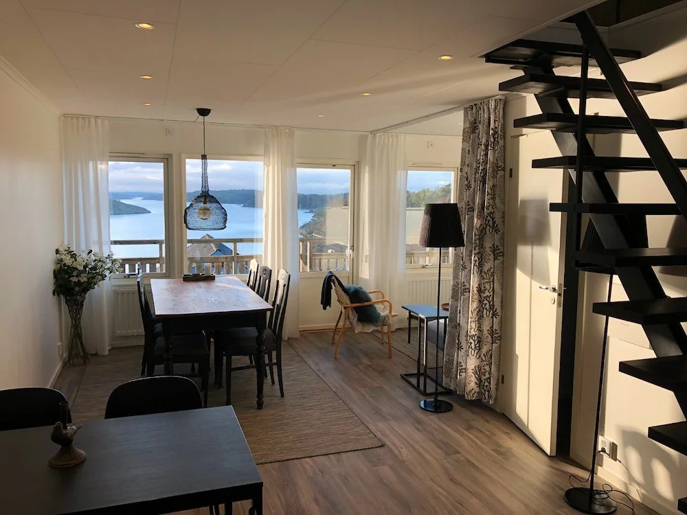 Vardagsrum med utsikt över sjön, matbord, stolar, gardiner, lampa, stol med filtar och skynke, trägolv, svart trappa.