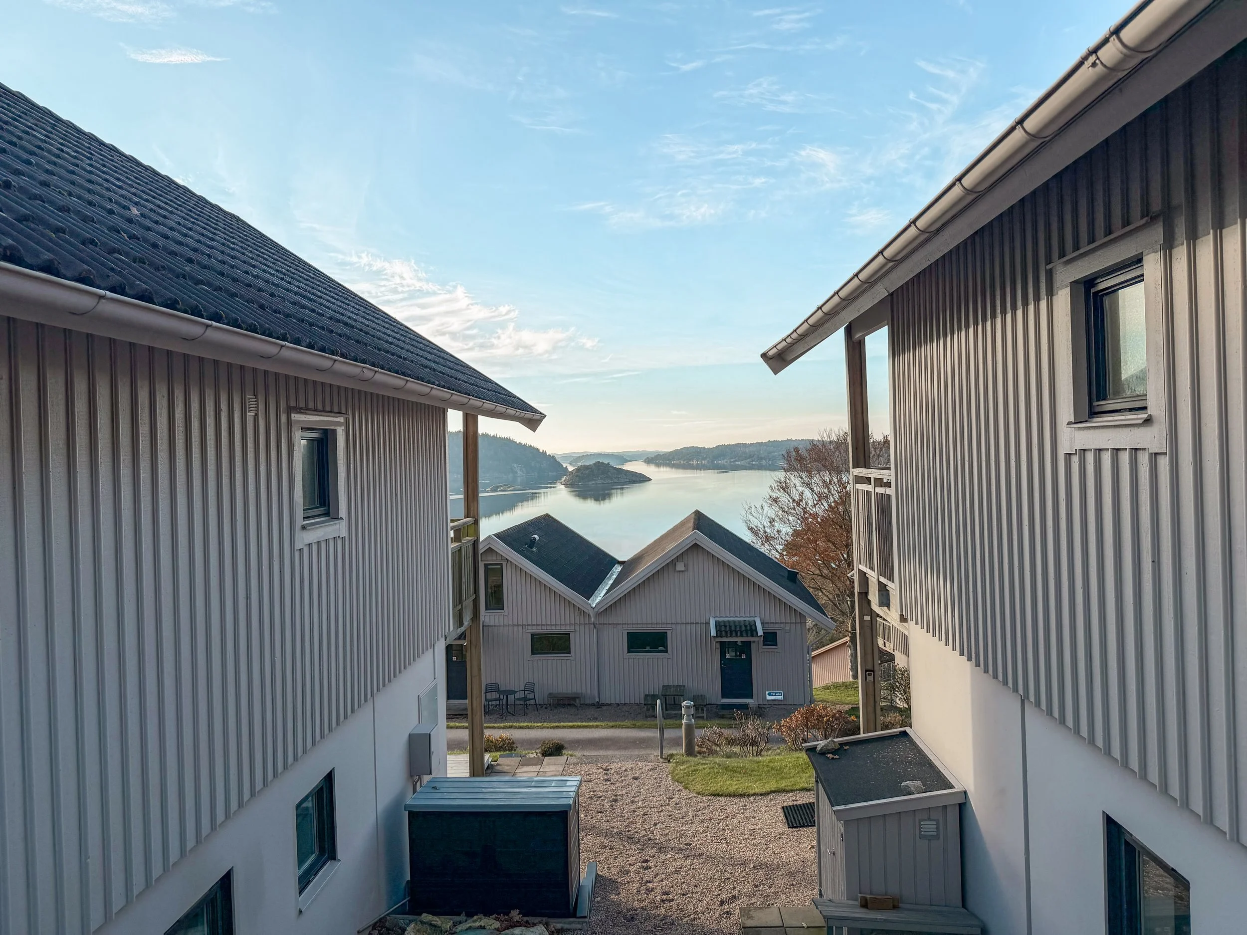 Två vita bostadshus med plåttak på vardera sida av en liten gångstig, utsikt över en vacker sjö och små öar i bakgrunden.