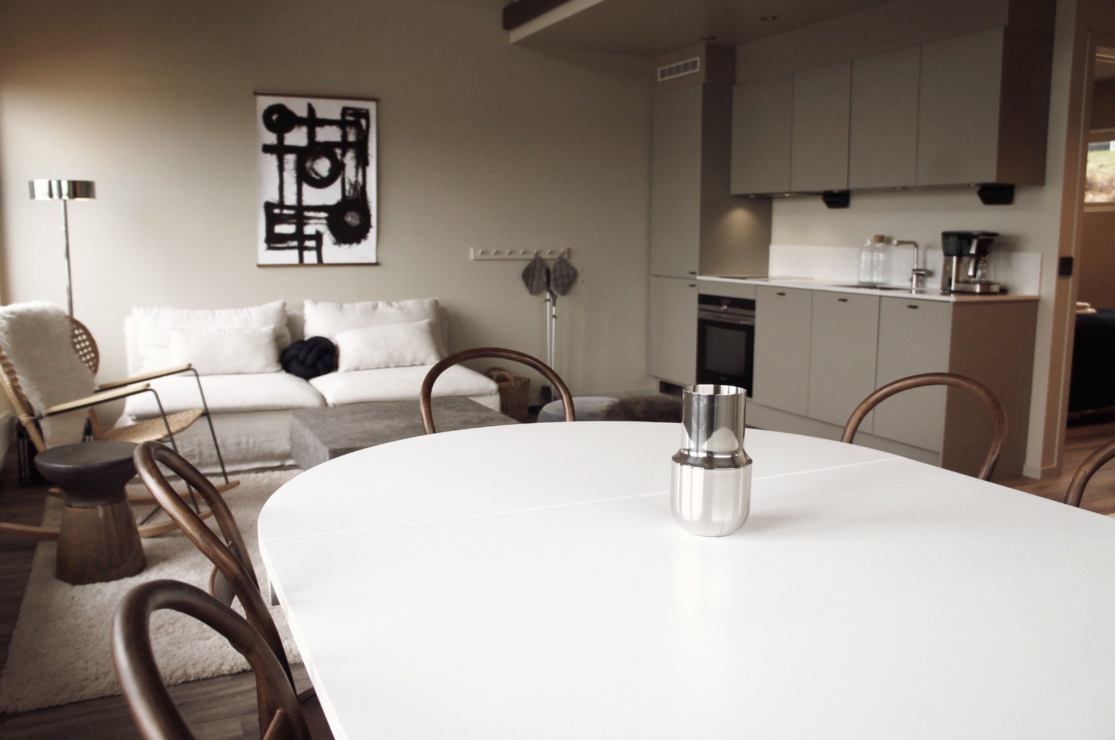 Vit matbord med tre trästolar i ett öppet kök och vardagsrum med soffa och abstrakt konst på vägg.