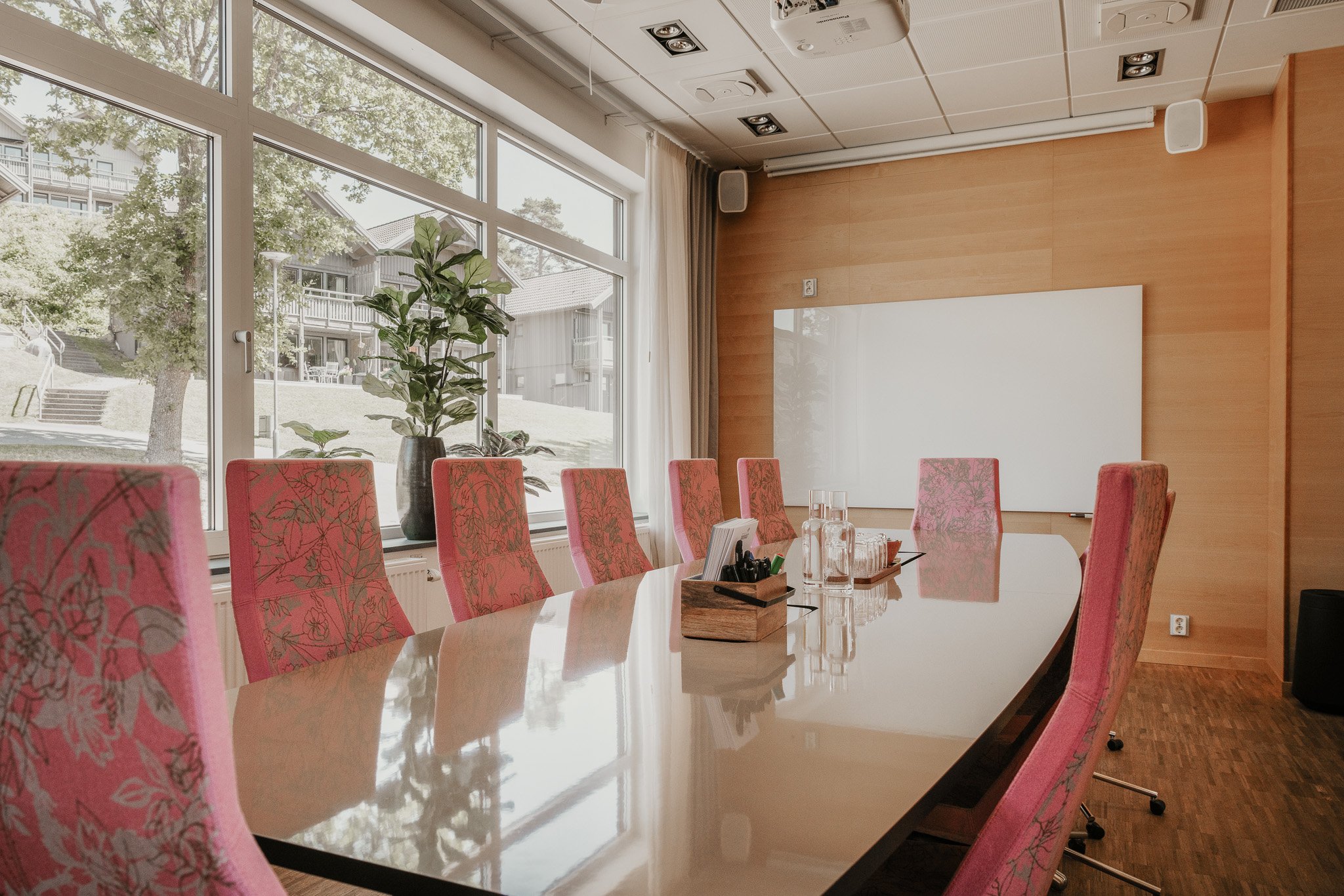 Salle de réunion avec une grande table blanche, des fauteuils roses à motifs de feuilles, de grandes fenêtres donnant sur des arbres et des bâtiments, une plante dans un vase, un tableau blanc au mur, des bouteilles en verre et des stylos sur la table.