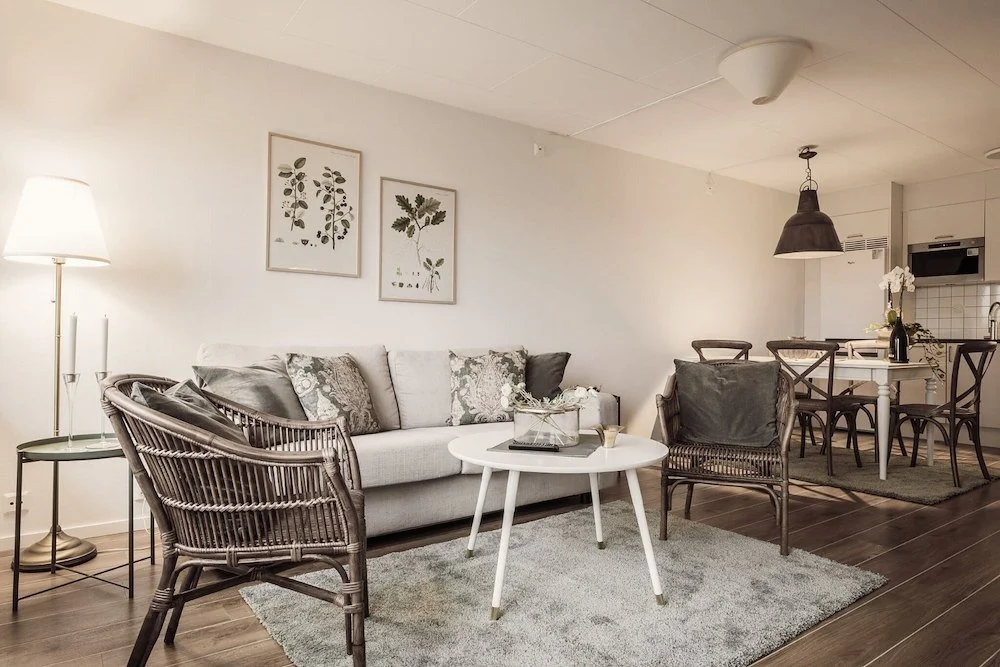 En inredningsbild av ett vardagsrum med en vit soffa, tre växtillustrationer på väggen, ett runt vitt soffbord med dekorationer, två korgstolar, en golvlampa, samt en del av ett kök med matbord och stolar i bakgrunden.