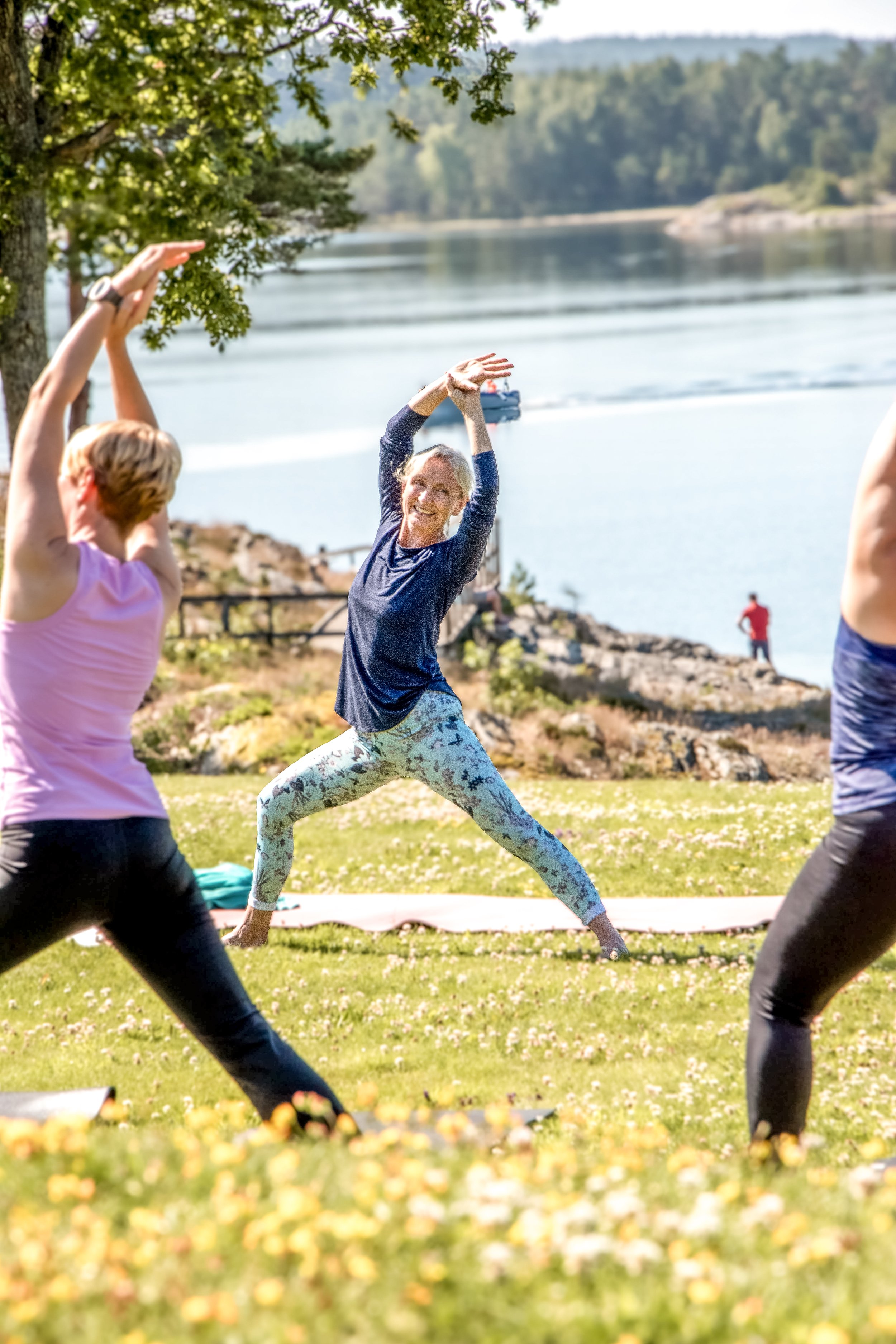 Flera personer utomhus som mediterar eller yoga på en gräsmatta precis vid havet, med träd och berg i bakgrunden, under en solig dag.