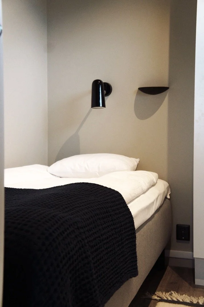Säng med vit lakan och svart filt, vägglampa och ett hörnskåp i ett litet sovrum.