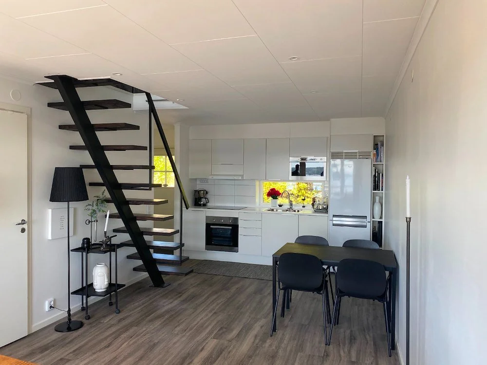 En modern, minimalistisk öppen planlösning med vit köksinredning, svart matbord med fyra stolar, ett svart trapphus, och ett hörn med bokhylla. Fönster som visar grönt landskap utanför.