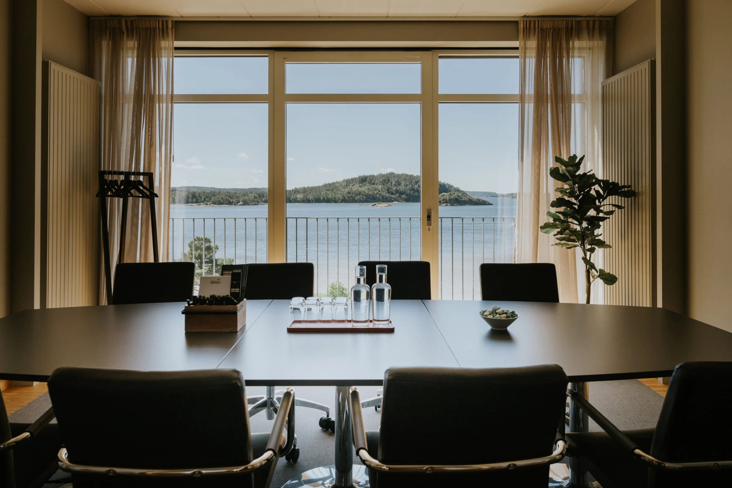 Konferensrum med stort fönster som visar en sjö och öar utomhus, inomhus finns ett bord, stolar, vattenkaraffer, glas, en växt och gardiner.