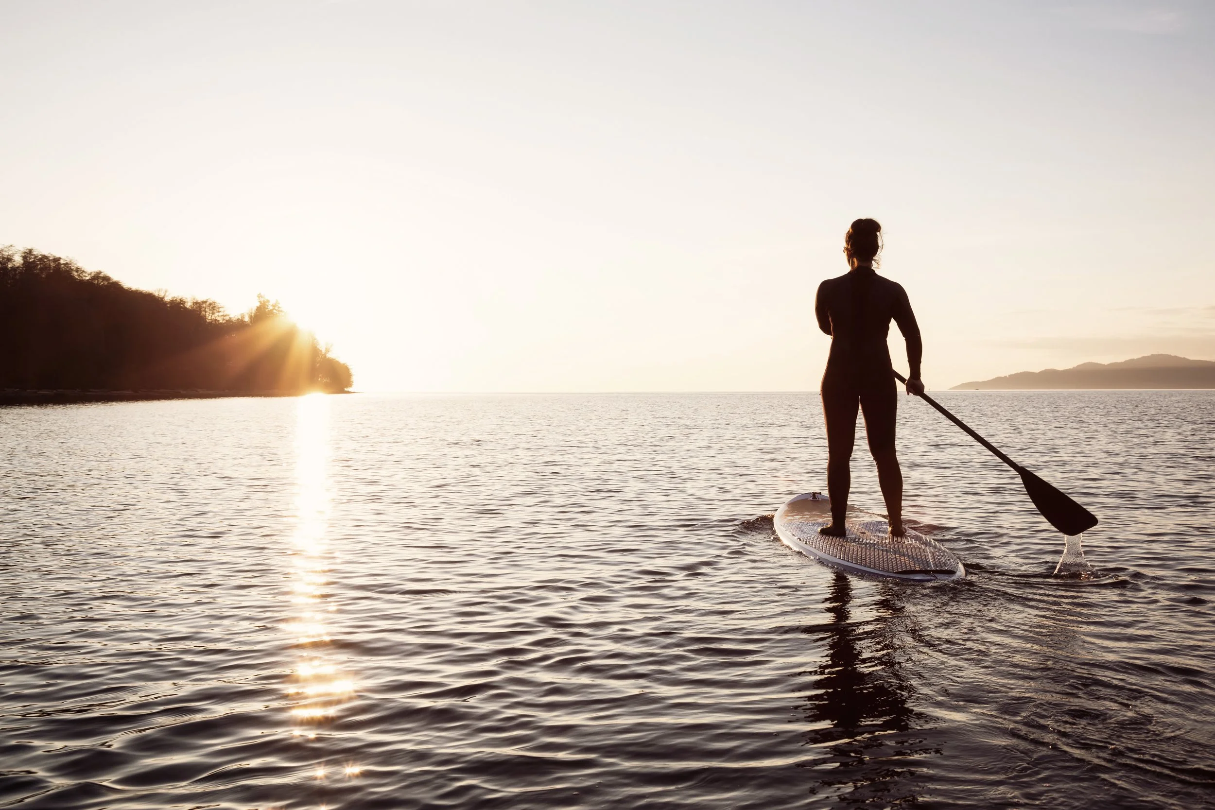 En person står på en stand-up paddleboard i vattnet mot solnedgången.