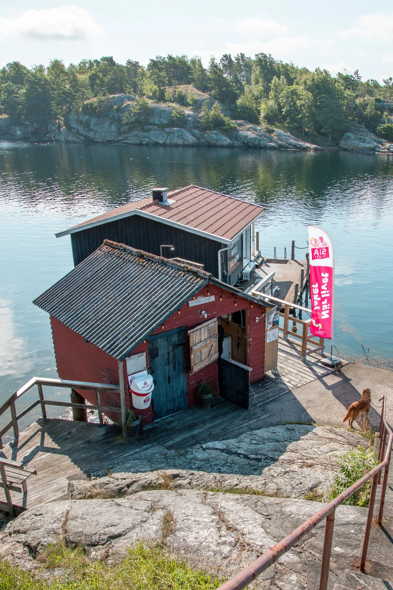 Två små trähus, ett rött och ett svart, vid vattnet med en brygga, på en klippa, med en hund som tittar ut mot vattnet. En logga och flagga med texten 'Wästgötbil' syns.
