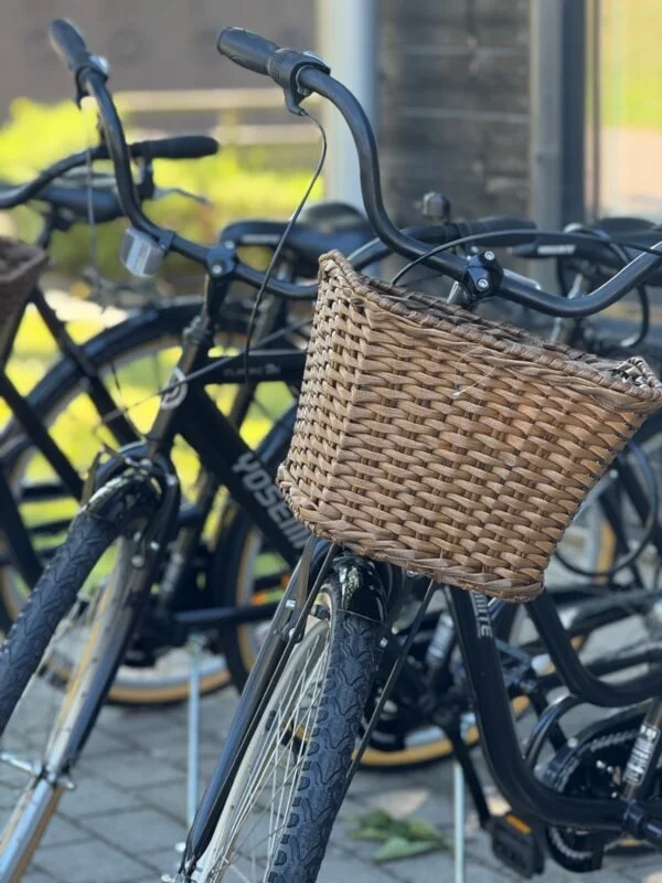 En svart cykel med en framtida korg av rottning på styret, parkerad bland andra cyklar utanför en byggnad.