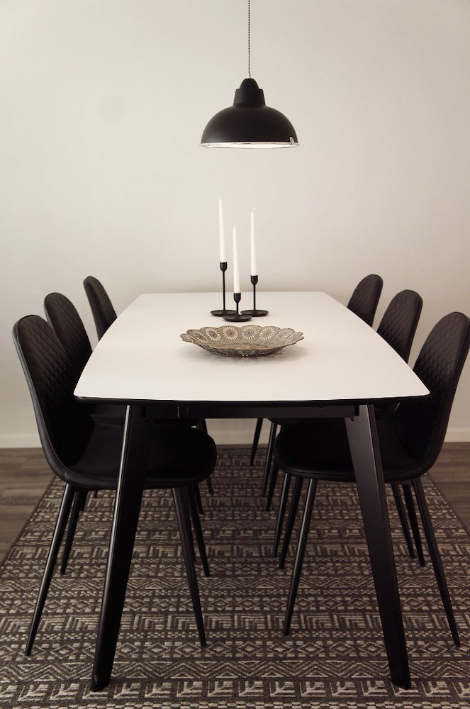 Svart taklampa hängande över vitt bord med svart stolar, ett metallfat och tre vita ljusstakar.