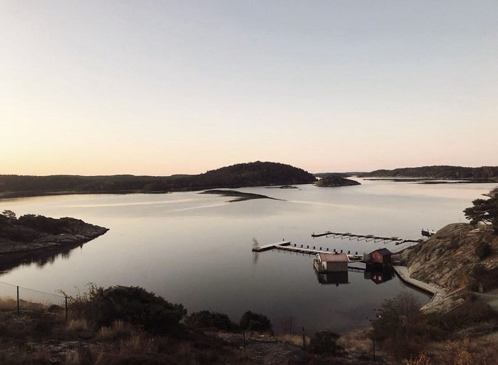 En lugn sjö med liten brygga och små hus vid vattnet, omgiven av klippor och ekar, vid skymning.