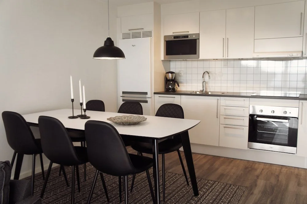 Kök med matbord och fyra svarta stolar, vita köksskåp, ugn, mikrovågsugn, kyl/frysskåp, kaffebryggare, och ett svart taklampor.