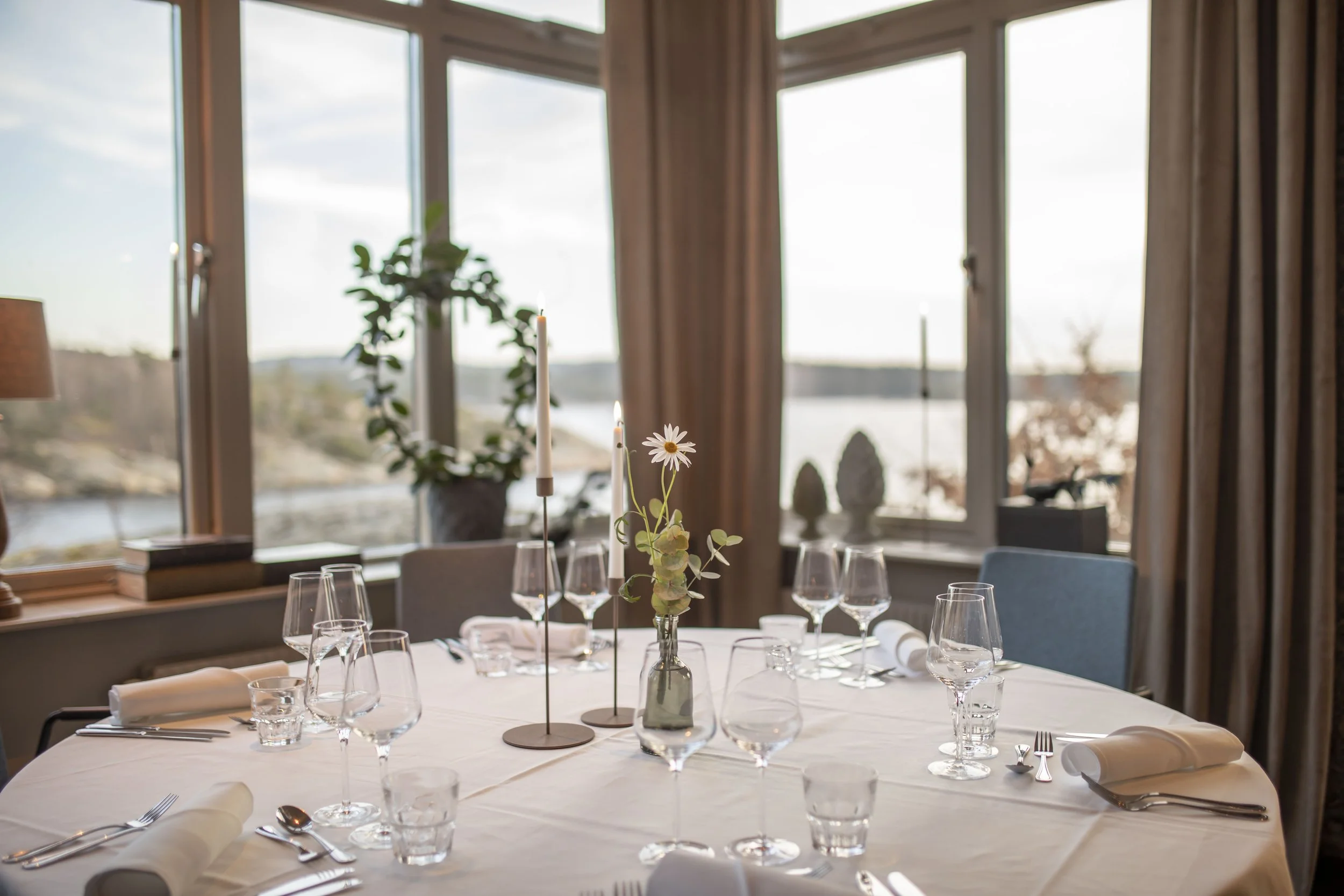 En formell middagsdukning på ett runt bord med vita servetter, glas, bestick och ljusstakar, placerade i ett rum med stora fönster och utsikt över vatten och landskap.