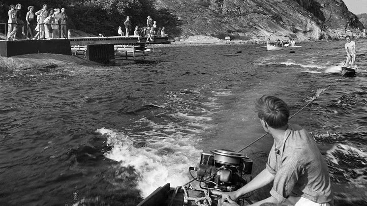 Män som åker motorbåt och fiskar i en sjö medan andra personer samlas på en brygga och tänger fisk.