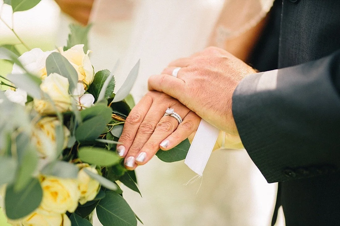 Närbild av ett brudpars händer med vigselringar, placerade på en blomsterbukett med gula och gröna blad.