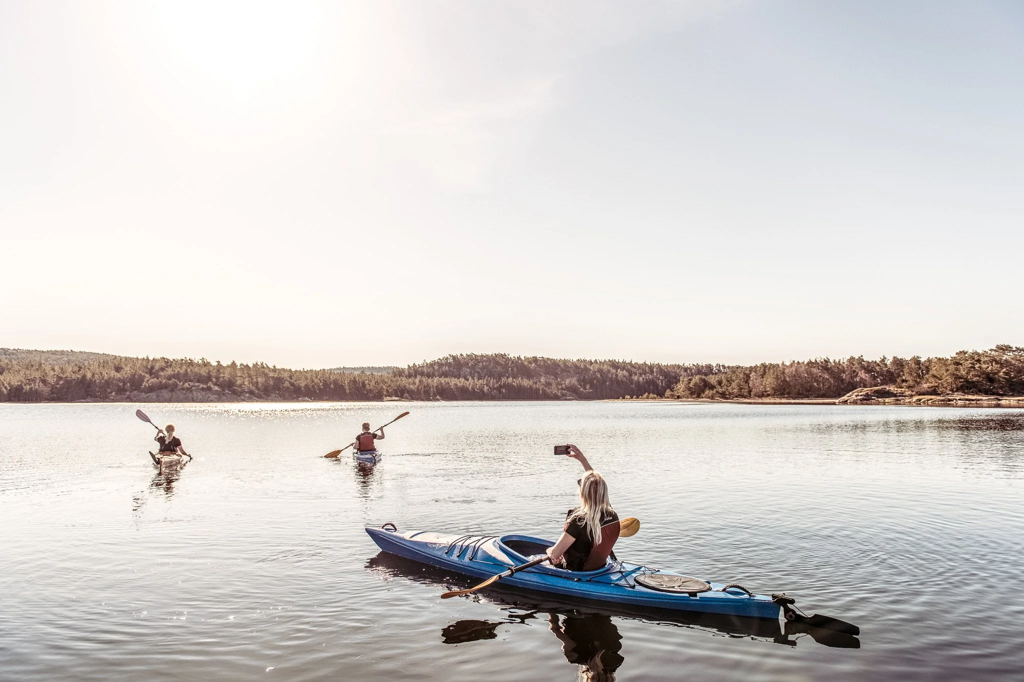 Tre personer i kajaker paddlar på en lugn sjö på en solig dag, med träd och skog i bakgrunden.