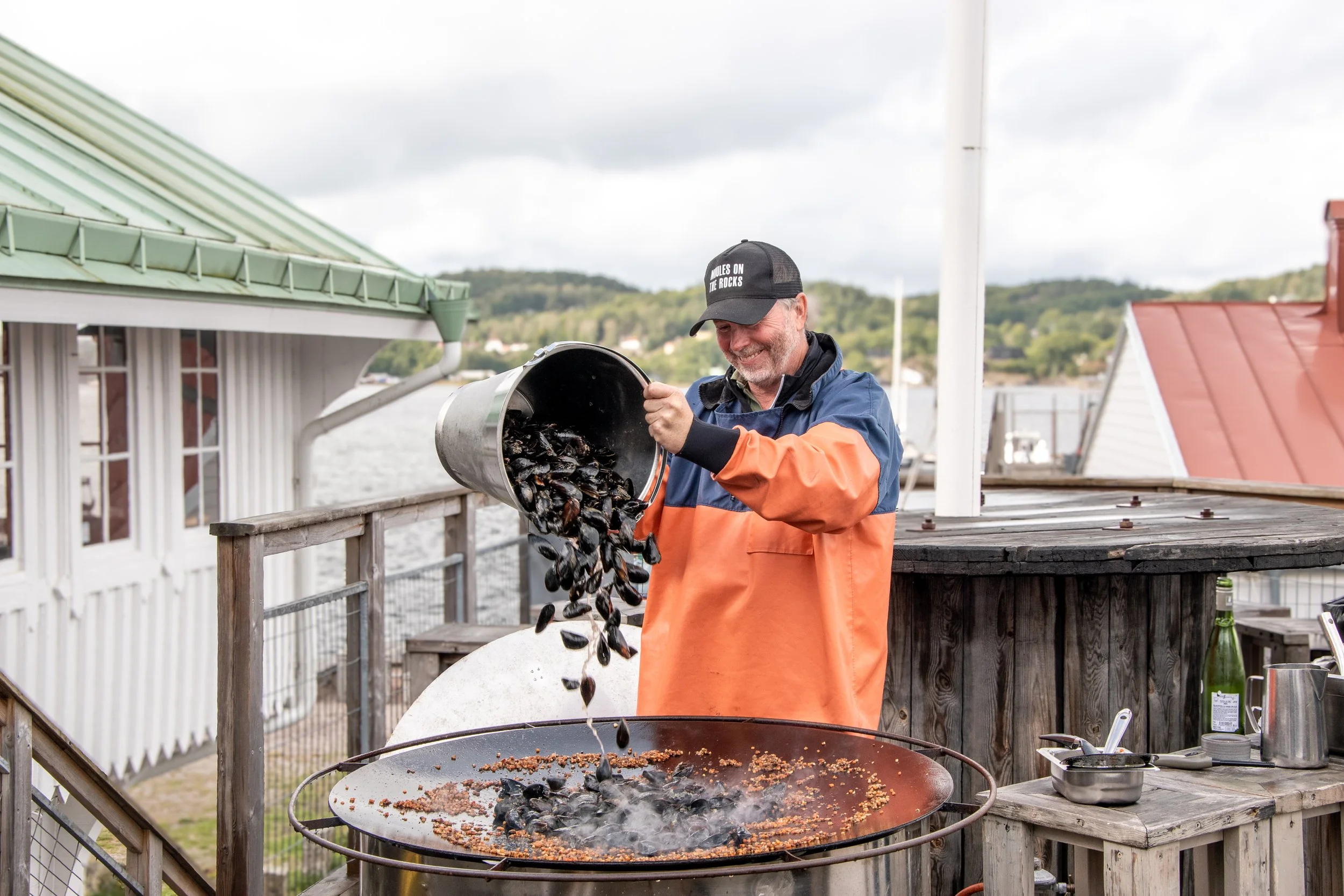 En man i en blå och orange jacka som grillar musslor på en öppen grill på en altan nära vattnet, med ett landskap av träd och hus i bakgrunden.