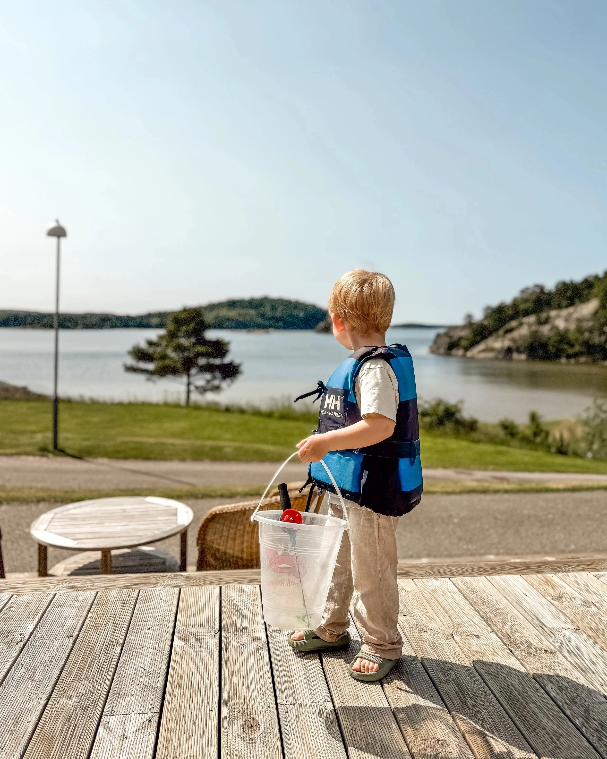 Ung pojke med flytväst som står på en uteplats, tittar ut mot vattnet med en hink och ett krabbfiskespö i handen.
