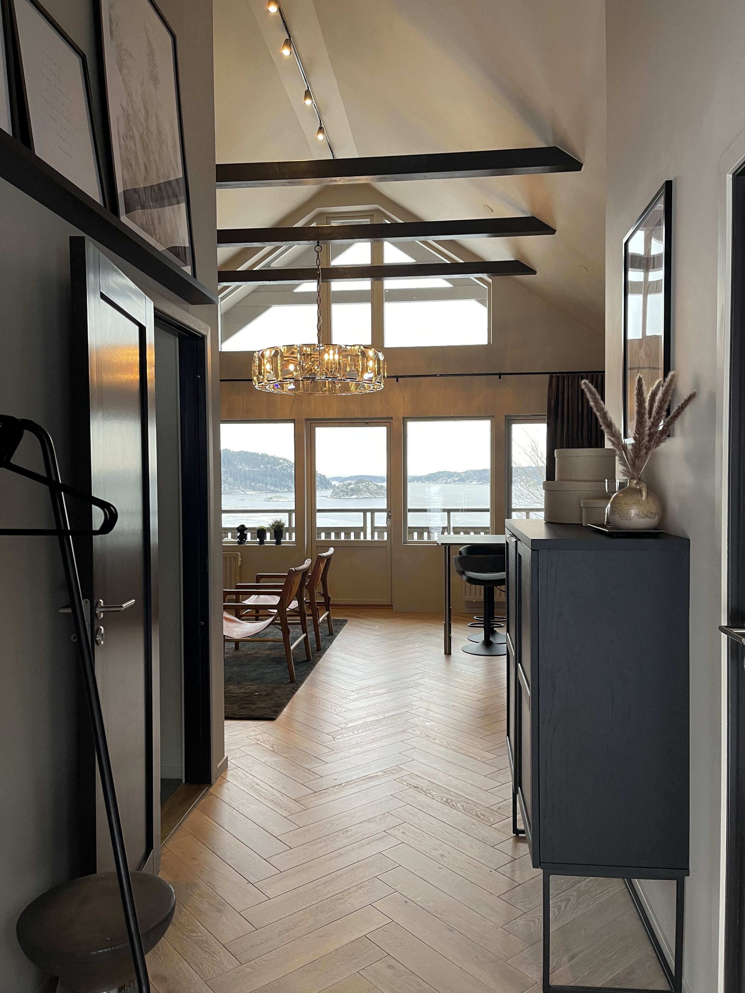 Bild av en ljus och modern semesterbostad med utsikt över vatten, stor fönstervägg, trädäck och trädgård utanför, stående ljuskrona, trägolv i fiskbensmönster, mörka möbler och dekorationer.