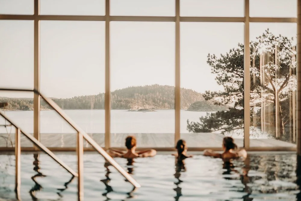 Tre personer kopplar av i ett poolområde med panoramafönster som visar en sjö och trädklädda kullar utomhus.