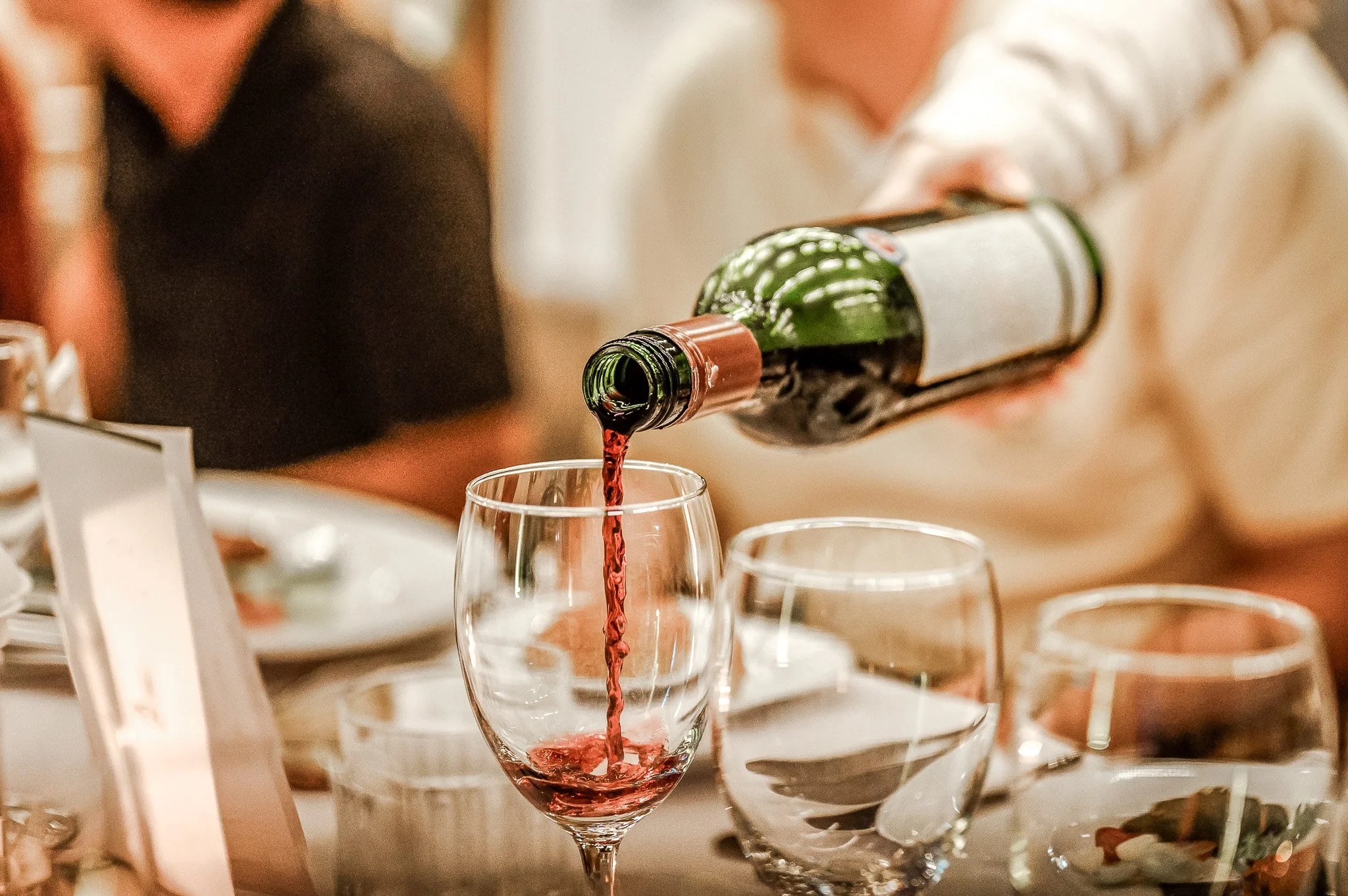 Person som häller rödvin i ett vinglas vid ett bord med andra glas och tallrikar.