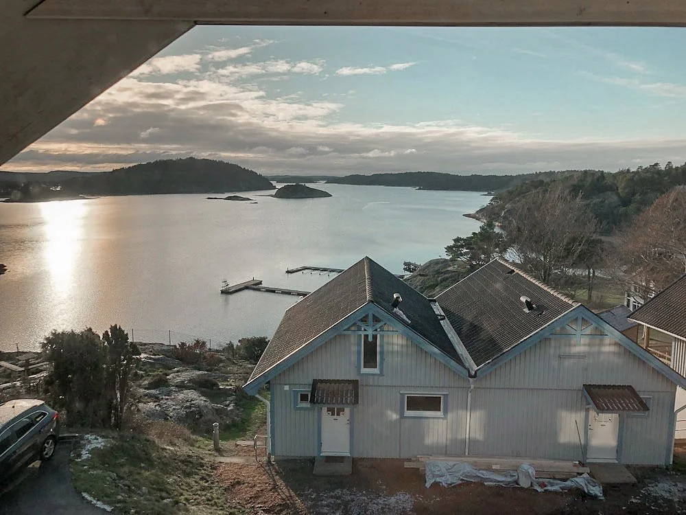 Utsikt över ett vattenlandskap med en liten trädhusliknande byggnad i förgrunden, omgiven av träd och en brygga på vattnet, under en molnig himmel.