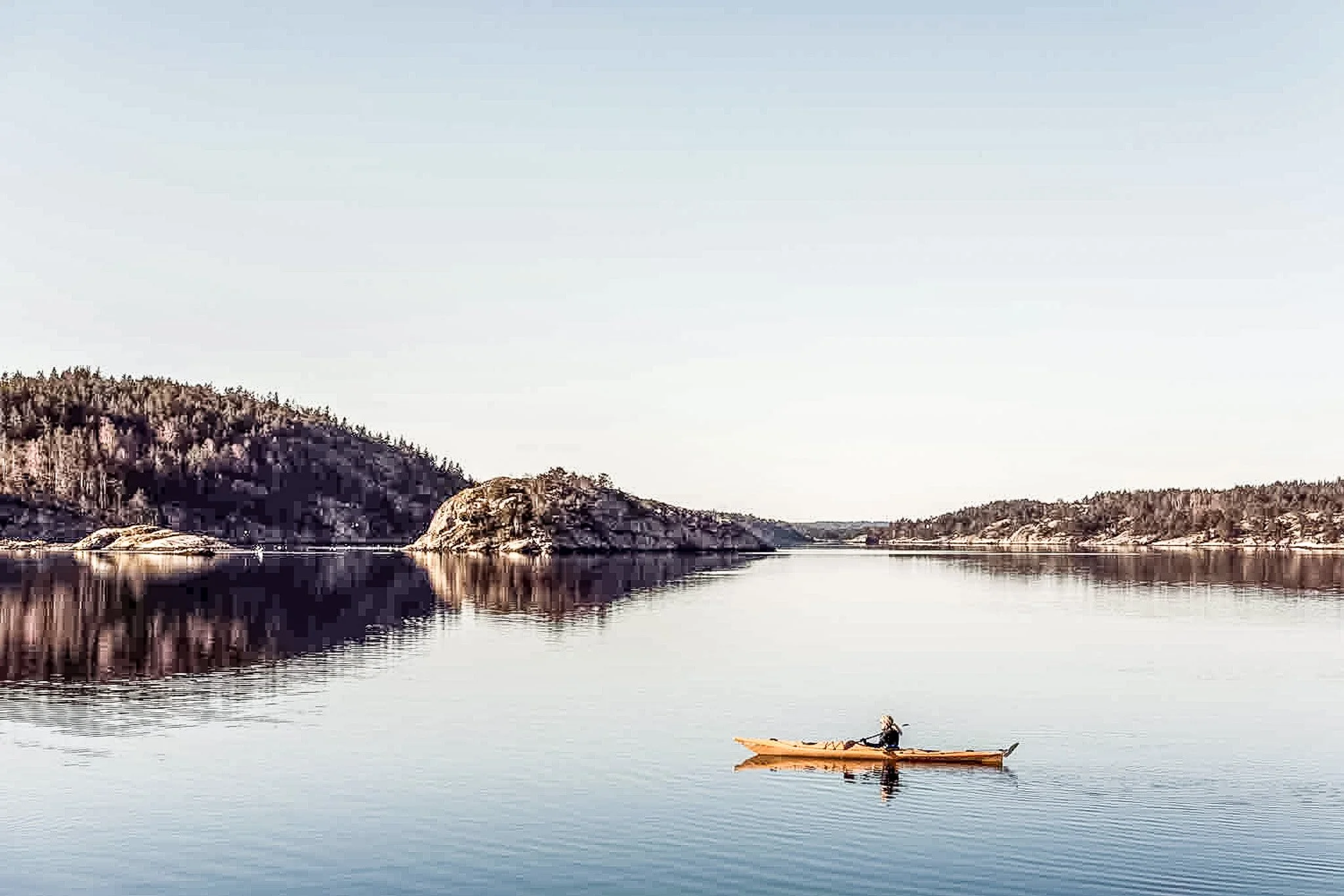En person paddlar i en kanot på en lugn flod omgiven av berg och skog.