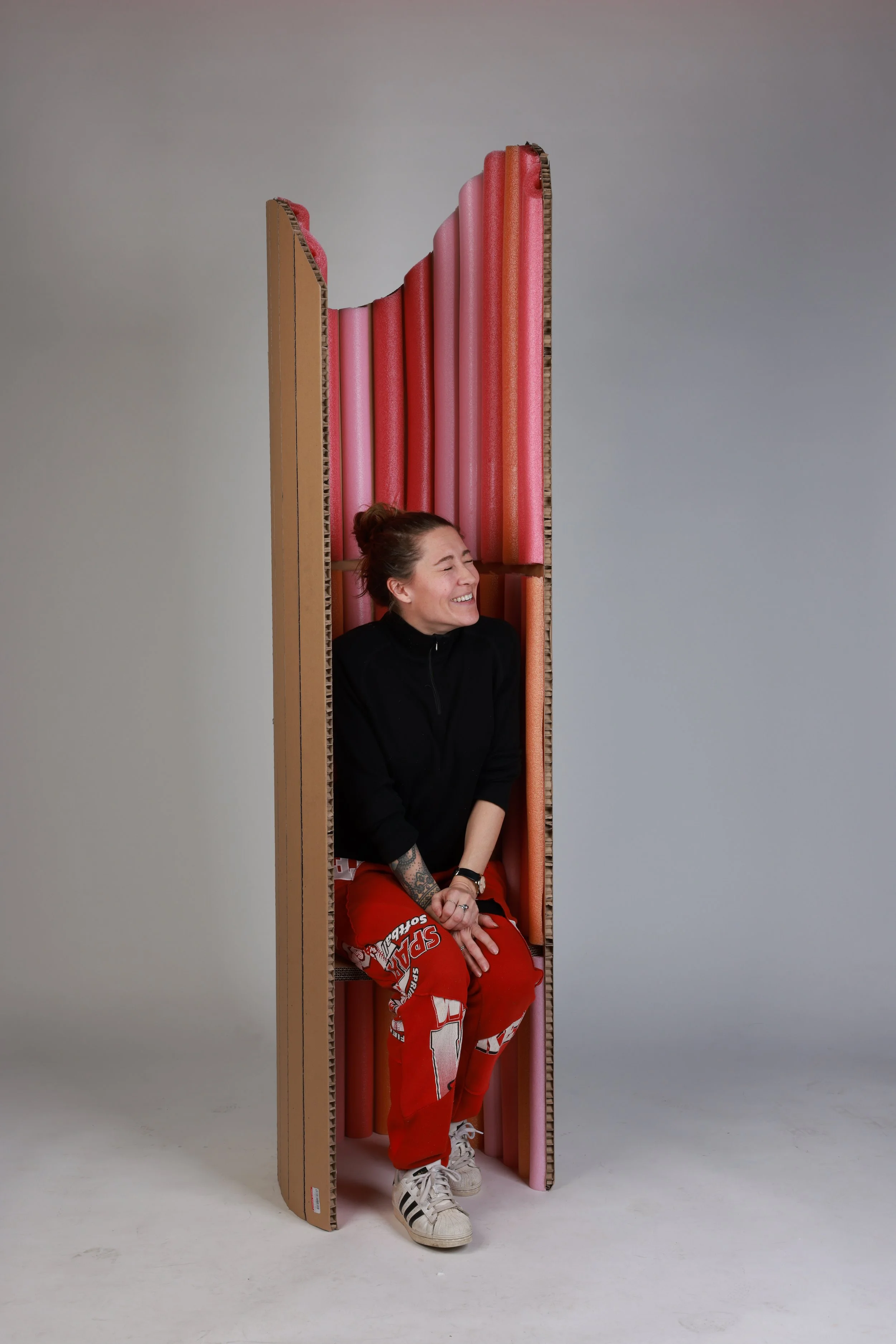 A woman sitting inside a cardboard structure filled with foam pool noodles, smiling and laughing, with light-colored background.