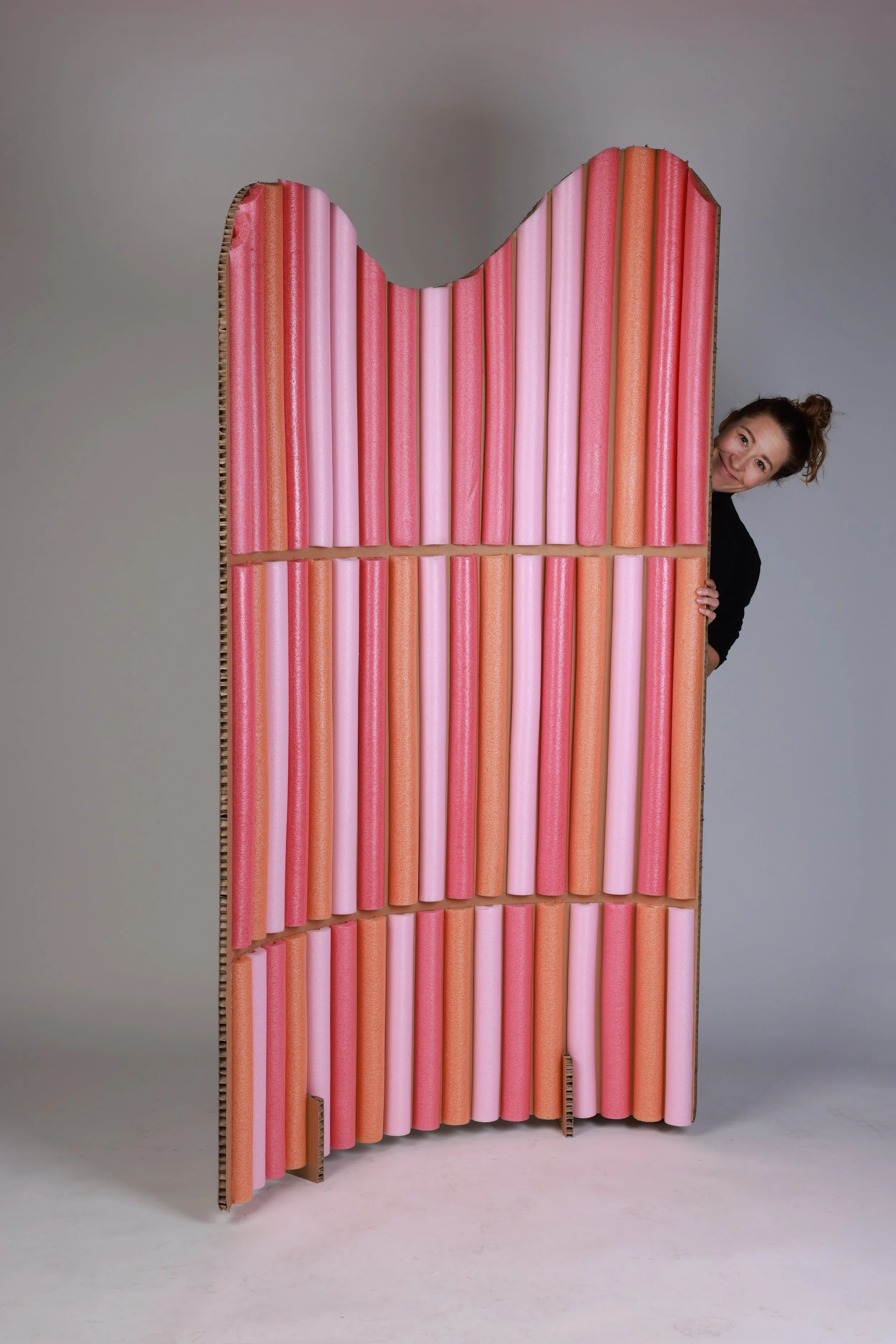 A woman peeking out from behind a large, colorful foam barrier with pink, red, and orange tubes arranged vertically.