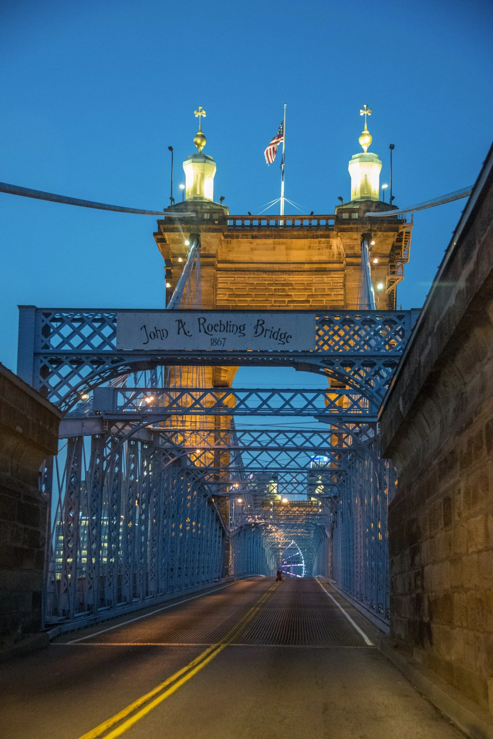 Nighttime view of the John A. Roebling Bridge, with the bridge's towers illuminated, American flag flying in the center. The road beneath is empty with yellow and white lines, framed by blue railings and brick walls.