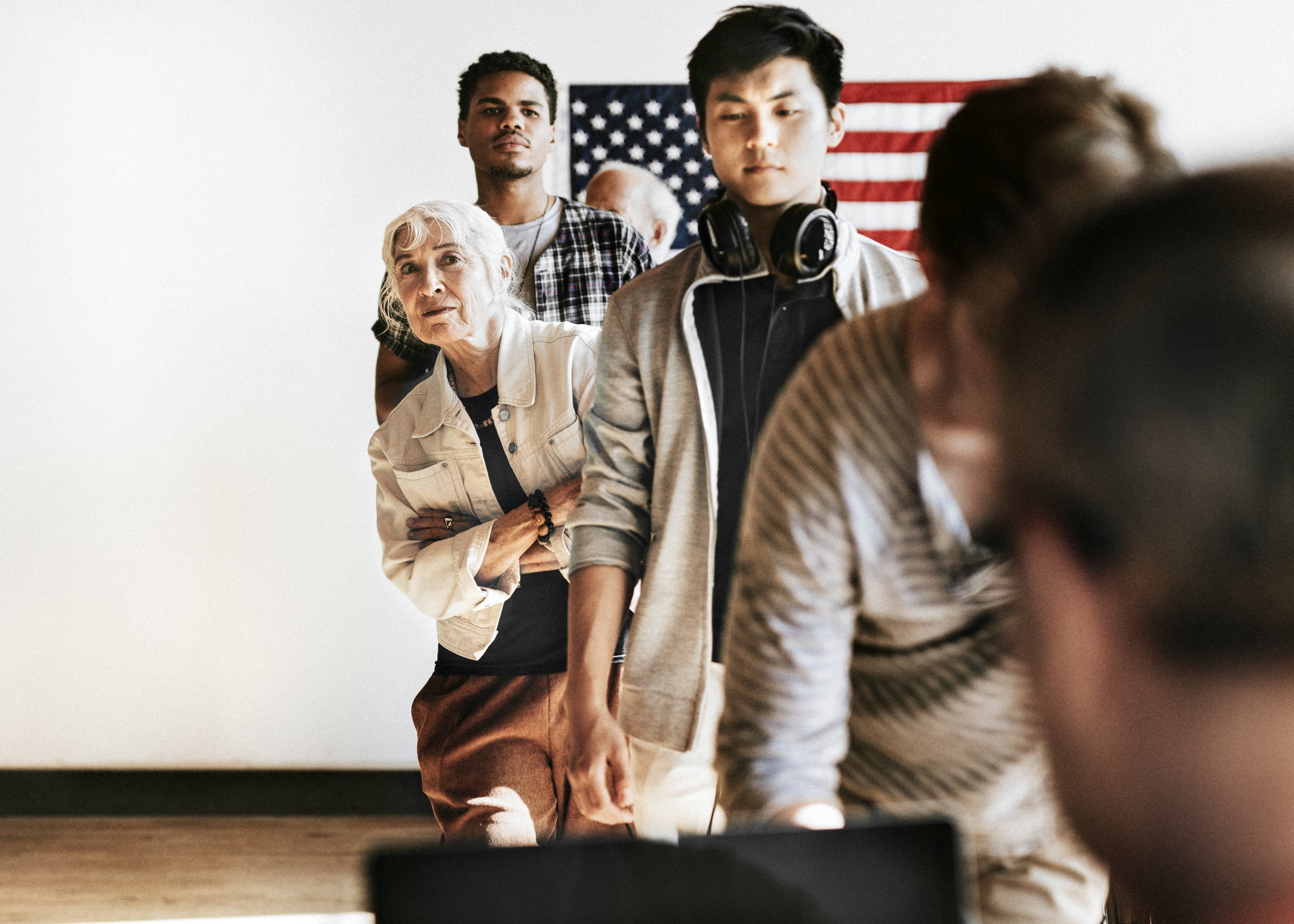 A group of people in line, with an American flag on the wall behind them, waiting or observing in an indoor setting.