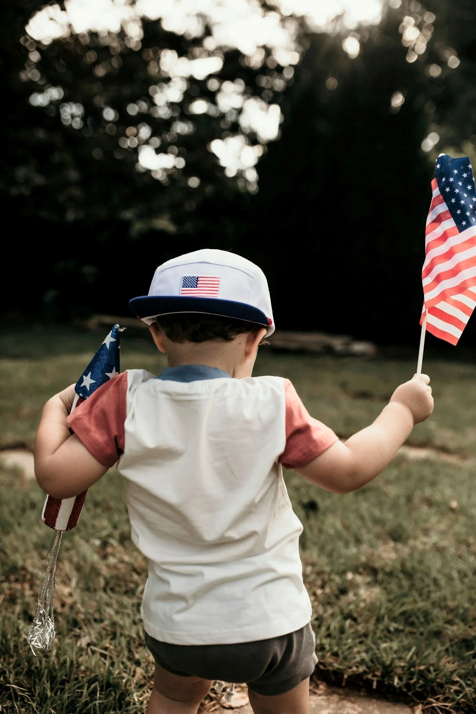 A young child wearing a white and blue cap with an American flag patch, holding American flags in both hands, standing outdoors on grass during the 4th of July with trees in the background.