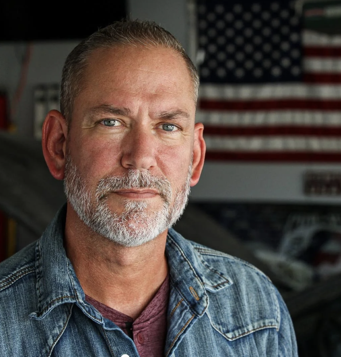 A middle-aged man with a gray beard and short hair, wearing a denim jacket and a maroon shirt, stands in front of an American flag displayed inside a garage or workshop.