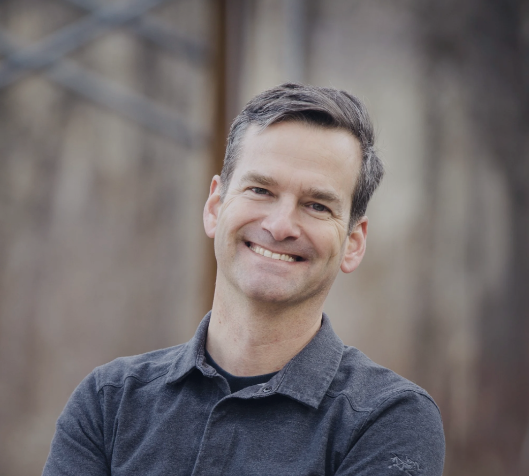 A smiling man with dark hair wearing a gray collared shirt outdoors, blurred natural background.