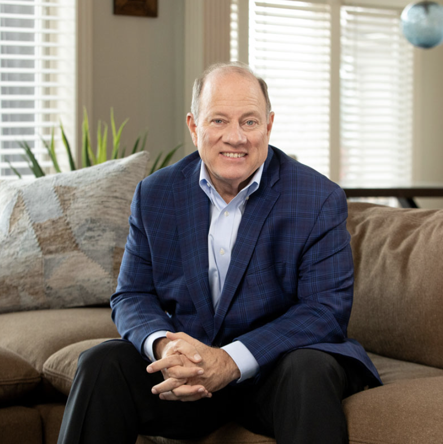 A middle-aged man in a blue checkered blazer and white shirt sitting on a beige sofa in a well-lit room with blinds and houseplants.