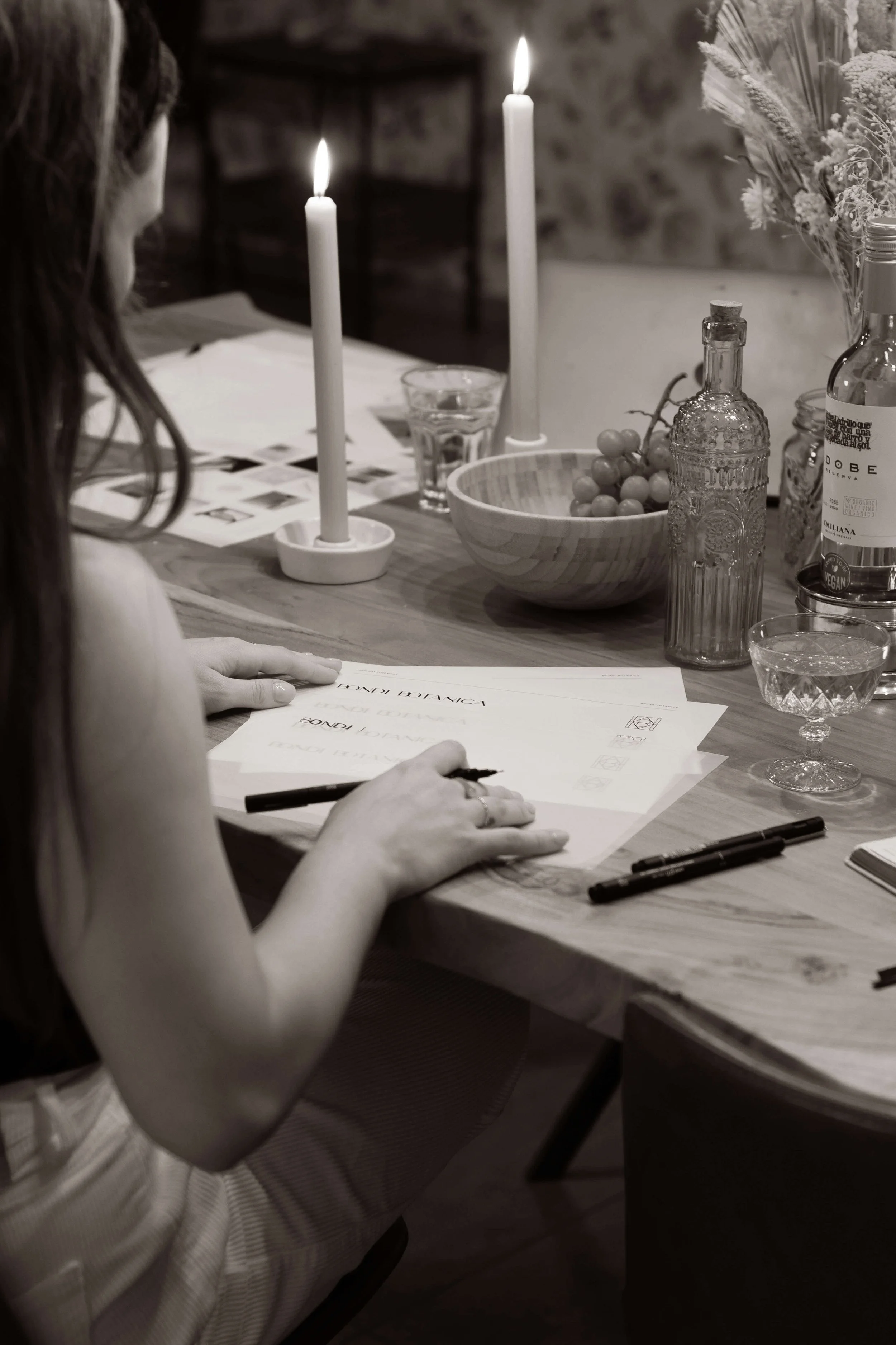 A person sitting at a dining table, writing on papers with candles, a bowl of grapes, and bottles on the table.