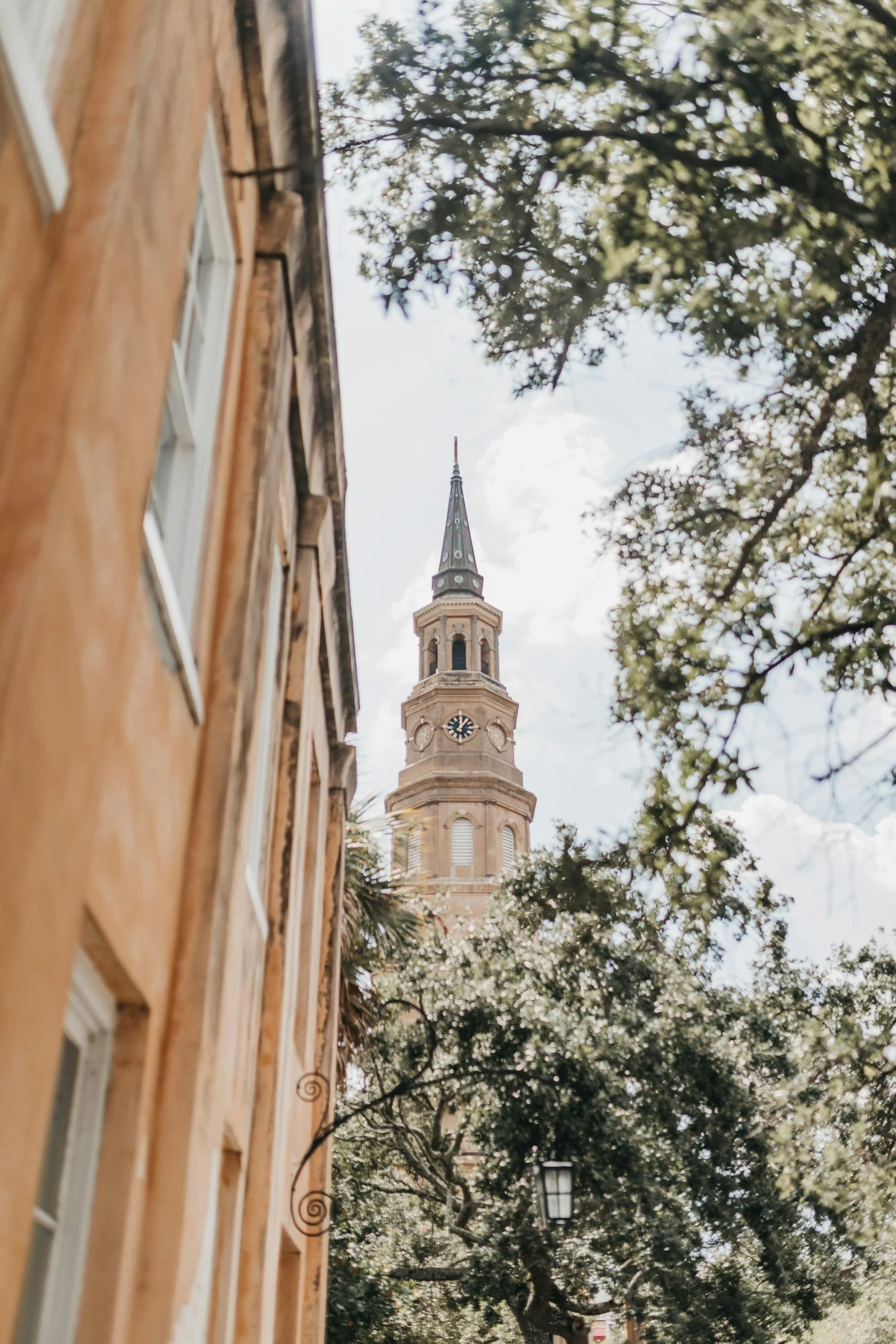View of a tall church steeple with a clock, seen through leafy trees and alongside old brick buildings.