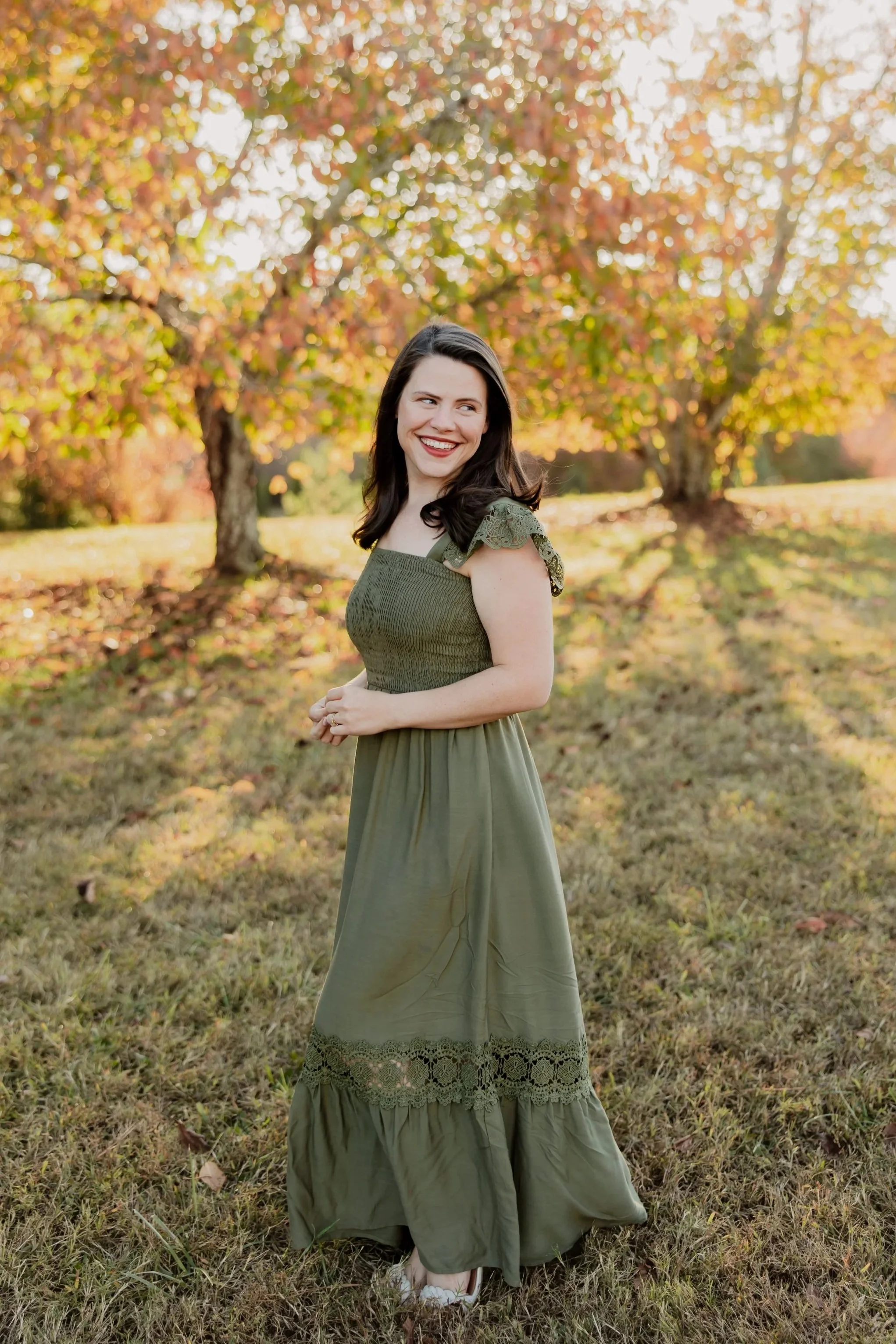 Woman, Founder of Bespoke Charleston, standing in a sunlit field during autumn, wearing an olive green dress with trees in the background.