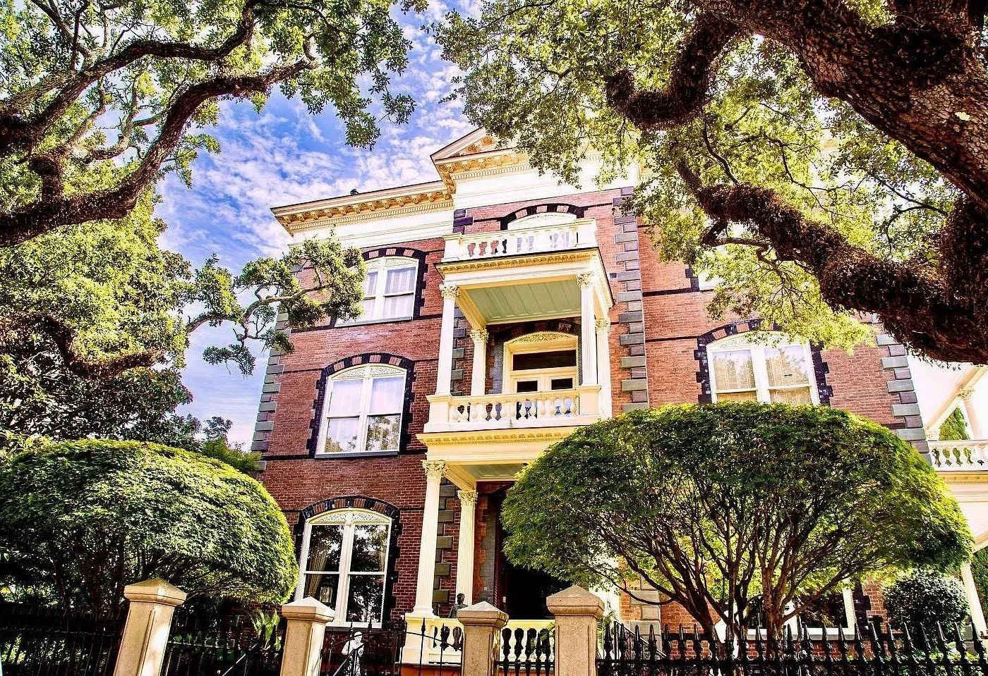 A large, historic Victorian house made of red brick with white accents, surrounded by large, leafy trees and lush bushes. The house features multiple balconies and arched windows with black trim.