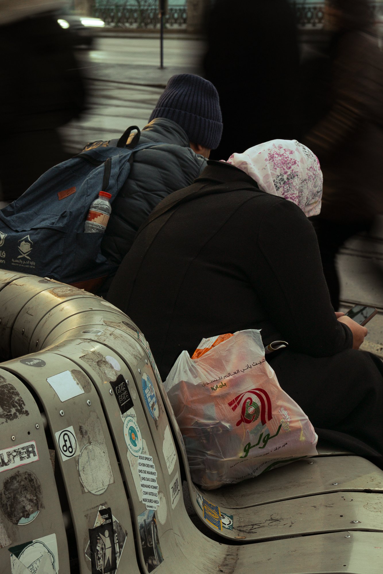 Two people sitting on a metal bench near subway tracks, one wearing a dark jacket and a blue knit cap, the other wearing a black hoodie and a white headscarf with floral pattern, with a backpack and a plastic bag nearby, alongside a row of coin-opera