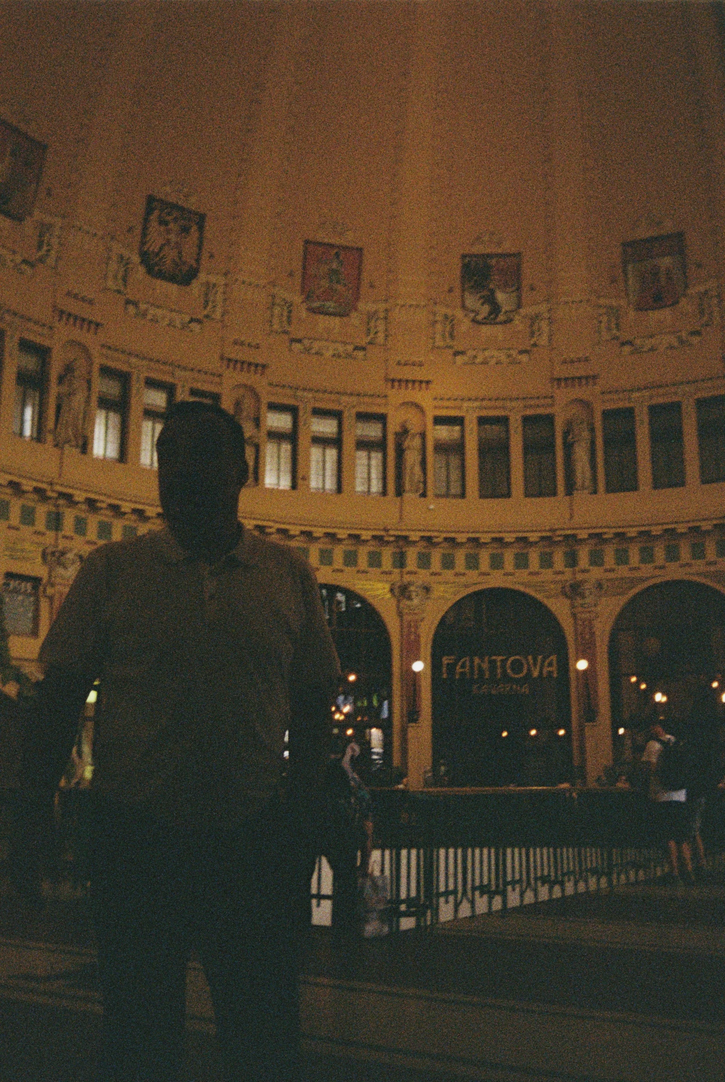 Nighttime view of a historic building with banners and statues, with the illuminated sign 'FANTOVA' and a person with a dark shirt in the foreground.